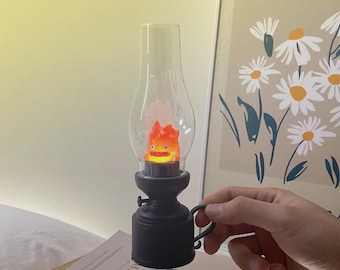 May include: Black and clear decorative lamp with a cartoon flame character inside. The lamp has a handle and sits on a book. The flame character is orange and yellow. The lamp is a decorative item.