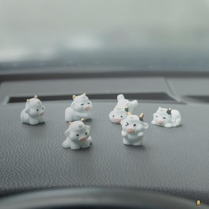May include: A collection of six small, white cow figurines with gold-colored horns and black markings. The miniature cows are in various poses, including sitting, lying down, and winking. The figurines are displayed on a dark gray surface.
