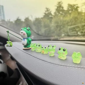 May include: A collection of green frog figurines, including a larger one with a blue scarf and a fishing rod with a small frog. The other frogs are small and translucent, with smiling faces. The figurines are arranged on a dark gray surface.