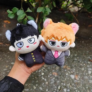 Puede incluir: Dos muñecos de peluche de personajes de anime. Uno tiene cabello negro y un uniforme oscuro, el otro tiene cabello naranja y un traje gris con corbata rosa. Ambos muñecos tienen ojos grandes y orejas de gato.