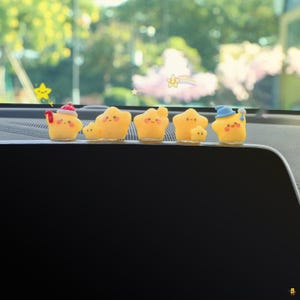 May include: A row of yellow star-shaped figurines with smiling faces and various small hats. The figurines are arranged on a dark surface, with a blurred background of green foliage and a bright sky.