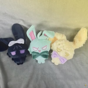 May include: Three plush bunny toys in dark blue, mint green, and yellow. Each bunny has a bow tie and a bow on its head. The bunnies have different facial expressions, with one appearing sleepy and another looking angry.