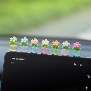 May include: A row of miniature flower pot decorations with colorful flowers. Each pot has a different colored flower, including pink, white, orange, and blue. The pots are clear with glitter inside. The background is a blurred green.