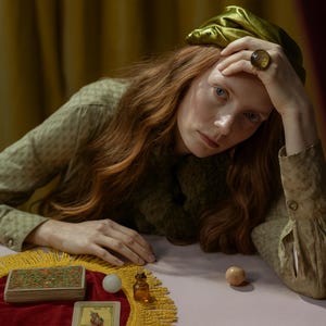 May include: A woman with long red hair wearing a green outfit and a gold ring with a large amber stone. A deck of tarot cards, a small bottle, and two spheres are on a table covered with a red cloth with a yellow fringe.