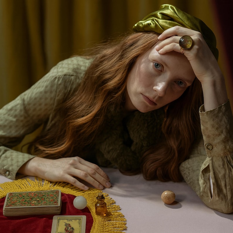 May include: A woman with long red hair wearing a green outfit and a gold ring with a large amber stone. A deck of tarot cards, a small bottle, and two spheres are on a table covered with a red cloth with a yellow fringe.