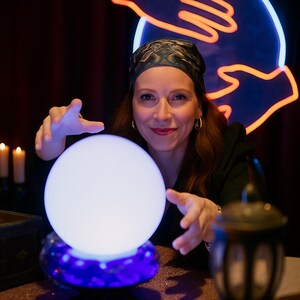 May include: A woman with long auburn hair and a patterned headscarf gazes at a glowing white crystal ball. The ball sits on a blue and purple base. The background includes a neon sign and candles.