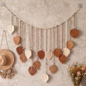 Peut inclure: Une tenture murale en macramé avec des ornements en forme de feuilles dans des tons de marron et de crème. Les feuilles sont suspendues à une ficelle horizontale avec des perles en bois. Un chapeau de paille et des fleurs séchées sont visibles en arrière-plan.