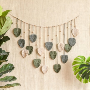 Puede incluir: Un tapiz de pared de macramé con diseños en forma de hojas en tonos verdes, grises y crema. Las hojas están suspendidas de una cuerda con cuentas de madera, creando una estética decorativa y natural. El fondo es beige neutro.