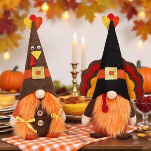 May include: Two Thanksgiving-themed gnome figurines. One gnome wears a brown hat with a yellow and red feather, and the other wears a black hat with a turkey feather design. Both have orange beards and are on a checkered tablecloth.
