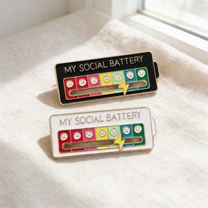 Puede incluir: Dos pines de esmalte con el texto "MY SOCIAL BATTERY". Un pin es negro con borde dorado, el otro es blanco con borde dorado. Ambos pines presentan un indicador de estilo batería con emoticonos y un rayo.