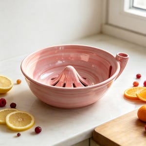 May include: A pink ceramic citrus juicer with a swirled marble pattern, featuring a central reamer cone, a bowl-shaped basin, and a small pouring spout on the side. The surface has a glossy finish with light and dark pink streaks throughout.