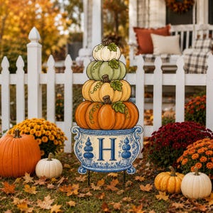 May include: A decorative yard sign featuring a stack of four pumpkins in white, light green, yellow, and orange, resting on a blue and white chinoiserie-style planter base with the letter H centered and two blue topiary illustrations on the sides.