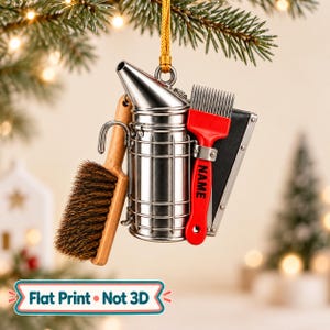 May include: A flat-printed hanging ornament featuring a silver metal bee smoker, a wooden-handled brush with brown bristles, and a red hive tool with the word NAME printed in black. The ornament hangs from a gold cord and includes a text label.
