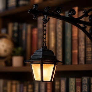 May include: A black metal hanging lantern with a tiered roof and frosted glass panels that emit a warm, glowing light. The lantern is suspended by a short black chain from a decorative, curved black metal wall bracket with scrollwork details.