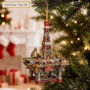 May include: A flat print Christmas tree ornament shaped like an oil rig with a red and silver derrick, a green helipad, and a sign that reads Merry Christmas. The structure is illuminated with small lights and hangs from a gold string.
