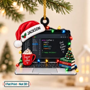 May include: A flat, laptop-shaped Christmas ornament featuring a red Santa hat with the name JACKSON, a screen displaying code, a red mug labeled FUELED BY COFFEE, and a stack of books labeled CODE, CREATE, BELONG, all wrapped in colorful lights.