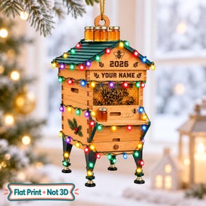 May include: A wooden beehive-shaped Christmas ornament featuring a green roof, three small honey jars on top, and a string of multicolored lights. The front displays the text 2026, YOUR NAME, and a bee graphic, with a visible honeycomb pattern.