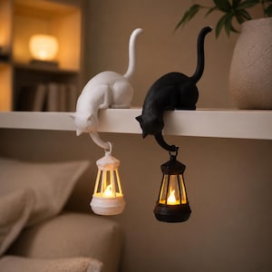 May include: Two cat figurines, one white and one black, perched on a ledge. Each cat has a long tail extending downward, holding a small lantern with a glowing, warm-toned light inside. The cats are positioned side-by-side on a flat surface.