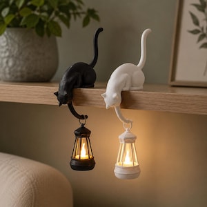 May include: Two cat-shaped ornaments, one black and one white, perched on a shelf edge. Each cat has a long tail extending downward, holding a small lantern with a glowing, flame-shaped light inside. The figures are smooth and solid in form.