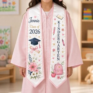 May include: A pink graduation gown with a white sash featuring the name "Jennie" and "Class of 2026." The sash is decorated with a mortarboard, diploma, and floral elements. The other sash says "Kindergarten" with school-themed illustrations.