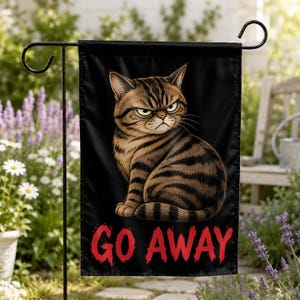 May include: A rectangular garden flag featuring a black background with a brown tabby cat displaying a grumpy facial expression. Below the cat, the text GO AWAY is printed in bold, distressed red capital letters. The flag hangs on a metal stand.
