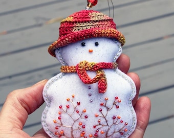 May include: A white felt snowman ornament featuring a crocheted red and gold hat, a matching crocheted scarf, and embroidered branches with small red and orange beads. The snowman has black bead eyes and a small orange nose, with a red bead topper.