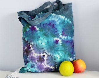 May include: A blue and purple tie-dye tote bag with two yellow apples in front of it. The bag has a long handle and is made of a soft, natural fabric.