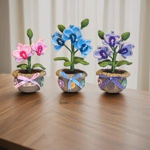 以下が含まれることがあります： 装飾的な鉢植えの、手作りのかぎ針編み蘭の植物が3つ。 蘭はピンク、青、紫の花と緑の茎と葉が特徴です。 灰色の鉢はリボンと装飾で飾られ、木製の表面に置かれています。