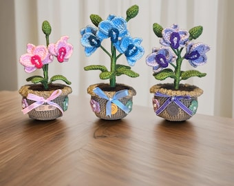 May include: Three handmade crochet orchid plants in decorative pots. The orchids feature pink, blue, and purple flowers with green stems and leaves. The pots are gray with decorative ribbons and embellishments, sitting on a wooden surface.