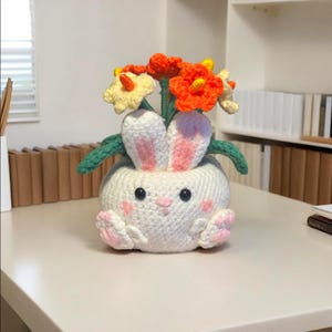 May include: A handmade, white crochet bunny planter with pink accents on the ears and paws. The bunny holds a bouquet of orange and yellow crochet flowers with green stems and leaves. The planter sits on a light-colored surface.