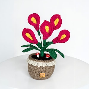 May include: A handcrafted crochet flower arrangement in a brown pot. The flowers are bright pink with yellow centres, green leaves, and two small mushroom decorations. The pot has a lace trim and a "MADE WITH LOVE" tag.