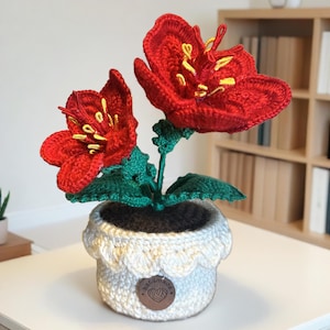 May include: A handmade crochet flower arrangement featuring two vibrant red flowers with yellow stamens, green leaves, and stems. The flowers are in a white crochet pot with a scalloped edge and a small, round tag that says "handmade".