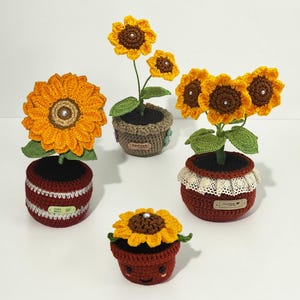 Puede incluir: Cuatro arreglos de girasoles de ganchillo en varias macetas. Tres tienen tallos altos con pétalos amarillos y centros marrones con cuentas blancas. Uno es una maceta pequeña, redonda y sonriente. Las macetas incluyen etiquetas Hand made.