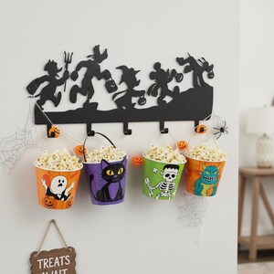 Może przedstawiać: Czarny, metalowy wieszak ścienny o tematyce Halloween z sylwetkami postaci i czterema wiaderkami popcornu. Wiaderka są pomarańczowe, fioletowe, zielone i pomarańczowe, każde z innym wzorem postaci z Halloween. Wiaderka są wypełnione popcornem.