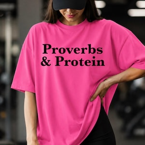 May include: A bright pink oversized t-shirt featuring the black text Proverbs & Protein printed on the chest. The shirt has short sleeves and a crew neckline, paired with black athletic shorts and a plain black baseball cap.