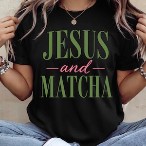 Puede incluir: Una camiseta negra de manga corta con el texto JESUS en letras verdes grandes, la palabra and en letra cursiva rosa más pequeña y la palabra MATCHA en letras verdes grandes. La prenda tiene un cuello redondo y es de tela negra lisa.