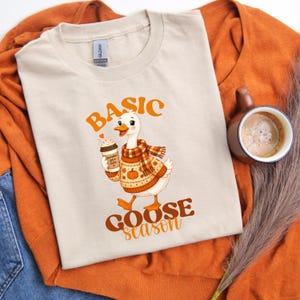May include: A beige crew neck t-shirt featuring a graphic of a white goose wearing a plaid scarf and an orange sweater with a pumpkin pattern. The goose holds a coffee cup labeled pumpkin spice latte. The text reads BASIC GOOSE season.