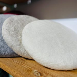 Puede incluir: Tres boinas de fieltro en tonos grises y blancos apiladas sobre una superficie de madera. Las boinas son redondas y tienen un aspecto suave y texturizado. Los colores van del gris oscuro al crema claro.