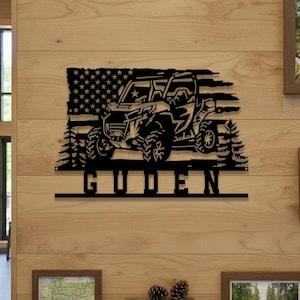 Op de afbeelding: Zwarte metalen wandkunst met een off-road voertuigontwerp, een Amerikaanse vlag als achtergrond en dennenbomen. De naam "GUDEN" staat eronder. Geschikt voor een garage of woonkamer.