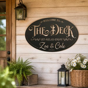 Può includere: Una targa da parete ovale nera con testo inciso di colore chiaro che recita WELCOME TO THE DECK, SIT RELAX ENJOY, Zoe & Cole, e EST.2023. La targa presenta decorazioni ornamentali ed è montata su una parete a doghe di legno chiaro.