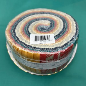 May include: A roll of fabric strips in various colours, including yellow, red, teal, and cream. The fabric is wrapped in clear plastic and sits on a white plate with a gold rim. The label reads "Valley-JR".