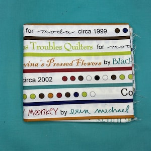 May include: Folded fabric with text including "for moda circa 1999", "Troubles Quilters", and "Monkey by erin michael". The fabric has a white background with colourful text and dots, and a brown and orange border.