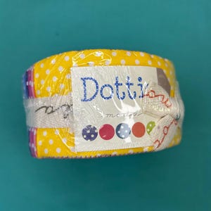 May include: A fabric roll wrapped in clear plastic. The fabric is yellow with white polka dots. A label reads "Dotti" in blue script. The label also features colourful dots and the word "moda".