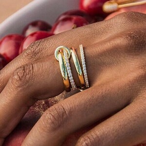 May include: A hand adorned with three gold rings, one with a knot design and two with small, clear stones. The hand is holding a partially cut grapefruit and is near a bowl of cherries.