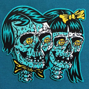 May include: A turquoise blue embroidered patch with two cartoon skulls with smiling faces. The skulls have yellow eyes with smiley faces inside. The skulls are wearing a bow tie and a yellow ribbon in their hair.