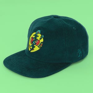 May include: A dark teal corduroy baseball cap featuring a central embroidered graphic of a yellow circular face split by a jagged blue lightning bolt with red eyes. A small matching embroidered character is visible on the side panel.
