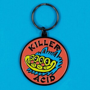 May include: A round keychain with a black ring and a red pendant. The pendant features a cartoon cat with blue fur, yellow teeth, and the words "KILLER ACID" in black.
