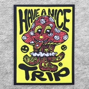 May include: A rectangular patch with a black border and a bright yellow background. The patch features a cartoon mushroom with a red cap and white spots, a smiling face, and the words "HAVE A NICE TRIP" in black lettering.