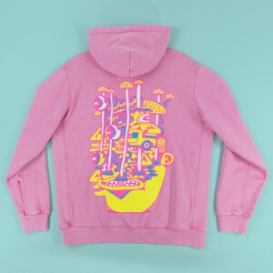 May include: Pink hoodie with a colorful graphic on the back. The graphic features a yellow face with a mushroom-like design in yellow, pink, and blue. The hoodie has a hood and long sleeves.