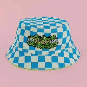 May include: A blue and white checkered bucket hat with a green and yellow embroidered alligator design. The hat has a green trim along the brim and is set against a pink background.